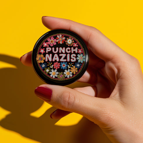 Round pinback metal button featuring an embroidered-style design on a black background. The center showcases bold, stitched-textured lettering in red, blue, and white that reads "PUNCH NAZIS." Surrounding the text is an intricate, folk-inspired floral pattern with multicolored flowers in shades of pink, orange, blue, and white, arranged symmetrically. A decorative zigzag border frames the edge of the button. The backside is silver metal with a secure pin closure.
