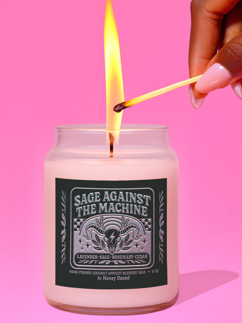 A 9-ounce, hand-poured scented candle in a transparent glass vessel. The candle contains a cotton wick and is filled with a creamy, off-white coconut apricot blended wax. It features a dark, matte finish label with a silver metallic emblem of a ram skull and stylized foliage, with the text "Sage Against The Machine" centered above the scent notes. A shiny gold lid sits atop the glass jar.