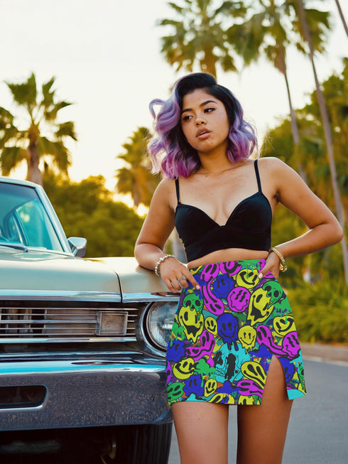 Vibrant mini skirt featuring an eye-popping psychedelic print of melting smiley faces and skulls in neon pink, bright yellow, electric blue, and lime green. The skirt has a distinctive side slit and sits above knee-length with a figure-hugging silhouette. Shown styled with various tops including a black crochet halter, black bustier, and purple blazer against colorful graffiti backdrops. The bold pattern makes this an unmissable statement piece with serious punk-meets-pop-art attitude.