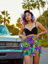 Vibrant mini skirt featuring an eye-popping psychedelic print of melting smiley faces and skulls in neon pink, bright yellow, electric blue, and lime green. The skirt has a distinctive side slit and sits above knee-length with a figure-hugging silhouette. Shown styled with various tops including a black crochet halter, black bustier, and purple blazer against colorful graffiti backdrops. The bold pattern makes this an unmissable statement piece with serious punk-meets-pop-art attitude.