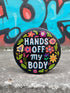 Glossy black pinback button with floral art and bold white text that reads “Hands Off My Body.” Feminist political button featuring bright pastel flowers in pink, red, orange, and yellow. Metal pin with durable safety pin back, shown in various settings: held by hand, displayed on pedestal, and against colorful street art. Available in 2" and 3" sizes, perfect for backpacks, jackets, tote bags, or as a gift for feminist allies and book lovers.