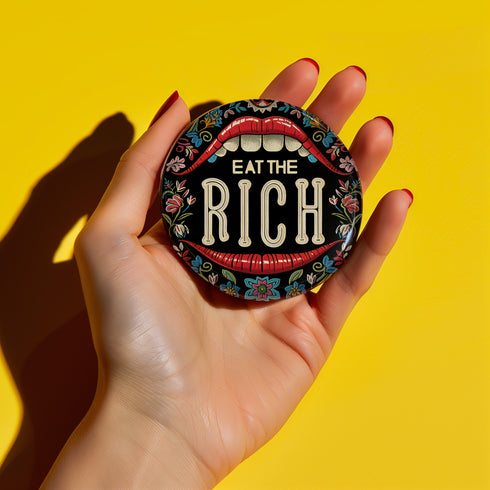 A round pin-back button with a bold, embroidered-style design featuring the phrase "EAT THE RICH" in large, vintage-style lettering. The typography is framed by a set of open red lips with detailed stitching, revealing white teeth. Surrounding the mouth are intricate floral patterns in shades of blue, red, and green, adding a decorative, folk-art-inspired touch. The button has a black background, making the details and lettering stand out