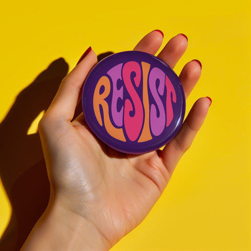 Close-up of a glossy, round purple pinback button. The button features the word "RESIST" in a bold, 70s-style psychedelic font that fills the entire surface. The bubbly, wavy letters alternate in color between a vibrant orange, a light peach-pink, and a deep magenta, creating a powerful and retro statement piece. The design has a distinct groovy and feminist protest aesthetic, making it a perfect accessory for jackets, bags, or activist apparel.