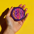 Close-up of a glossy, round purple pinback button. The button features the word "RESIST" in a bold, 70s-style psychedelic font that fills the entire surface. The bubbly, wavy letters alternate in color between a vibrant orange, a light peach-pink, and a deep magenta, creating a powerful and retro statement piece. The design has a distinct groovy and feminist protest aesthetic, making it a perfect accessory for jackets, bags, or activist apparel.