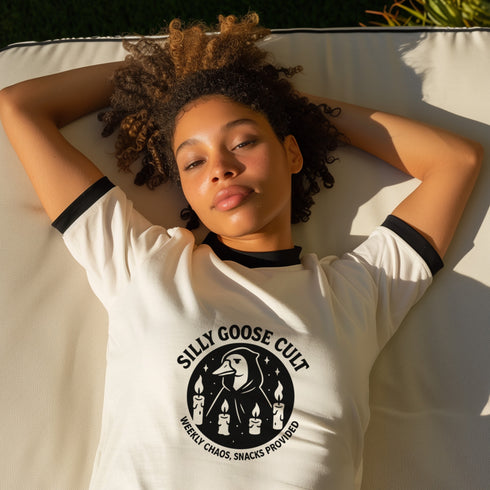 Unisex ringer t-shirt in natural cotton with black ribbed collar and sleeve cuffs. Front features bold black graphic: a cult-style goose surrounded by four candles, with text reading “Silly Goose Cult, Weekly Chaos, Snacks Provided.” Available in sizes S to 3XL. Retro-inspired fit, soft cotton, statement weirdcore design for alternative fashion lovers.