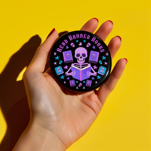 Literary rebel's badge of honor. Black circular button featuring a neon purple skeleton surrounded by floating books and stars. The text 'READ BANNED BOOKS' arcs across the top in bold purple lettering. Perfect for book-loving misfits who devour forbidden knowledge.