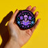 Literary rebel's badge of honor. Black circular button featuring a neon purple skeleton surrounded by floating books and stars. The text 'READ BANNED BOOKS' arcs across the top in bold purple lettering. Perfect for book-loving misfits who devour forbidden knowledge.