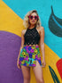 Vibrant mini skirt featuring an eye-popping psychedelic print of melting smiley faces and skulls in neon pink, bright yellow, electric blue, and lime green. The skirt has a distinctive side slit and sits above knee-length with a figure-hugging silhouette. Shown styled with various tops including a black crochet halter, black bustier, and purple blazer against colorful graffiti backdrops. The bold pattern makes this an unmissable statement piece with serious punk-meets-pop-art attitude.