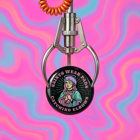 Pinback button badge featuring a saint-inspired roller skater wearing pads and a helmet, with pink hair and teal robes. Surrounded by retro starbursts, the black button reads “Saints Wear Pads Catching Elbows” in pastel pink and teal lettering. Design is bold, colorful, and printed on a glossy metal surface with a classic safety pin back. Available in 2.25" or 3" sizes.