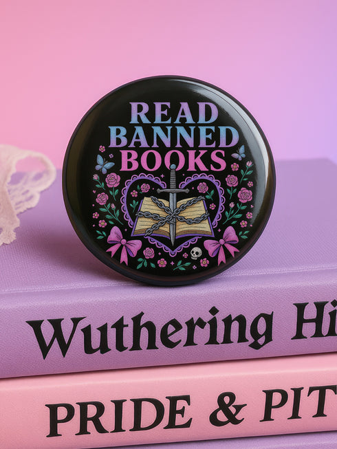 Glossy black pinback button that says “Read Banned Books” in purple and teal letters, featuring a chained book, roses, bows, and floral accents, styled on colorful books and pastel backgrounds.