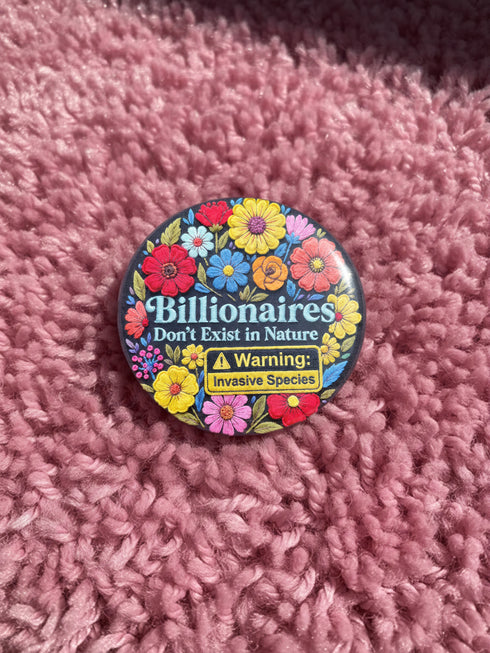 A round pinback button featuring a flat-printed design that mimics the appearance of highly detailed, 3D embroidery (trompe-l'œil). The background is solid black, with a floral border including red, yellow, sky blue, magenta, and orange flowers. In the center, the text "Billionaires Don't Exist in Nature" is written in a blue serif font. Below the text is a yellow rectangle with black borders containing a warning icon and the words "Warning: Invasive Species" in a bold, sans-serif black font.