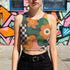 Cropped racerback tank top featuring bold retro floral print with orange and yellow mod flowers, green leaves, black and white checkerboard pattern, and abstract wavy designs. Fitted athletic style crop top in vibrant 70s inspired botanical print with performance fabric construction. Women's festival rave wear cropped camisole with psychedelic flower power design perfect for summer concerts, Y2K fashion looks, and alternative streetwear styling.