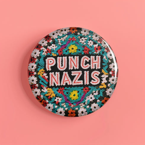 Round pinback metal button with an embroidered-style design on a black background. The center displays the phrase "PUNCH NAZIS" in bold, white stitched-textured lettering with red accents. Surrounding the text is an elaborate folk-inspired floral pattern, densely packed with embroidered-style flowers in shades of red, yellow, blue, pink, and white. The intricate design includes symmetrical lace-like stitching and decorative motifs, creating a rich, tapestry-like effect.