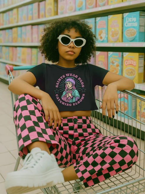 Black cropped t-shirt featuring a colorful, stylized saint figure with pink hair, wearing a helmet and pads. Surrounded by radiant lines and the phrase “Saints Wear Pads Catching Elbows” in bold pastel text. The design has a vintage punk-meets-holy aesthetic with star accents and a retro vibe. Ideal for roller skate girls, derby athletes, and goth skater fans. Available in sizes XS–2XL with raw hem details.