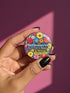 A round pinback button featuring a flat-printed design that mimics the appearance of highly detailed, 3D embroidery (trompe-l'œil). The background is solid black, with a floral border including red, yellow, sky blue, magenta, and orange flowers. In the center, the text "Billionaires Don't Exist in Nature" is written in a blue serif font. Below the text is a yellow rectangle with black borders containing a warning icon and the words "Warning: Invasive Species" in a bold, sans-serif black font.