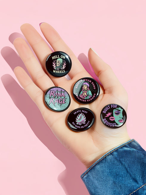 A set of five round 2.25-inch pinback buttons featuring original roller derby designs including a bruised face, saint on skates, flaming roller skate, skull in helmet, and bold text phrases like “Built for Bruises” and “Hell on Wheels.” Each button has a glossy black finish with vibrant pastel pink, purple, mint, and aqua artwork, mounted on metal tinplate with a safety pin back.