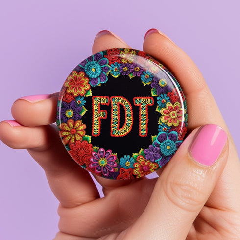 Circular black pinback button with bright 'FDT' lettering in orange/teal surrounded by colorful embroidery-style flowers in red, purple, blue, and yellow. Metal safety pin backing visible on reverse side. Political protest pin with vibrant Mexican folk art-inspired floral design.