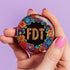 Circular black pinback button with bright 'FDT' lettering in orange/teal surrounded by colorful embroidery-style flowers in red, purple, blue, and yellow. Metal safety pin backing visible on reverse side. Political protest pin with vibrant Mexican folk art-inspired floral design.