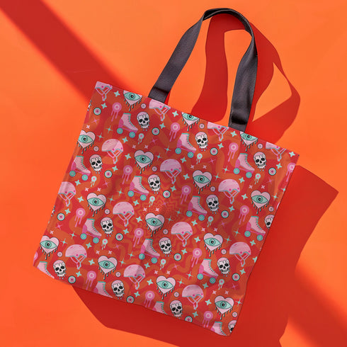 Tote bag with black handles featuring a vibrant red background covered in pastel pink and blue illustrations of skulls, roller skates, eyes, and geometric shapes. Displayed on an orange surface with dramatic shadows.