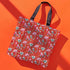 Tote bag with black handles featuring a vibrant red background covered in pastel pink and blue illustrations of skulls, roller skates, eyes, and geometric shapes. Displayed on an orange surface with dramatic shadows.
