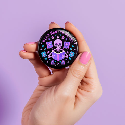 Literary rebel's badge of honor. Black circular button featuring a neon purple skeleton surrounded by floating books and stars. The text 'READ BANNED BOOKS' arcs across the top in bold purple lettering. Perfect for book-loving misfits who devour forbidden knowledge.