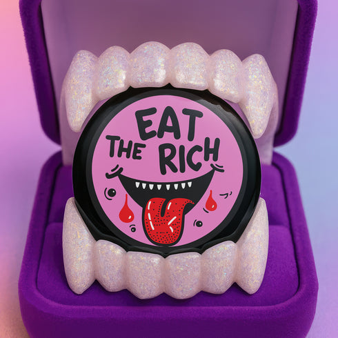 Round pink pinback button featuring a cartoon-style open mouth with sharp teeth, a red tongue, and dripping red details. The bold black text reads “EAT THE RICH” in a hand-drawn style. Glossy black rim with a vibrant, rebellious graphic design.