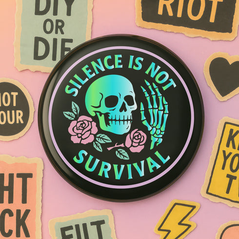 Round enamel pinback button available in 2.5″ or 3″ sizes with a black base and high-gloss finish. The design features neon green and blue gradient skull in the center, pastel pink roses and green leaves below, and mint skeleton hand on the right side. Arched text reads “SILENCE IS NOT SURVIVAL” in teal block letters. Shown styled on a denim jacket, held in hand against bright yellow backdrop, placed in a candy heart flat lay, and propped on pavement in front of graffiti wall.