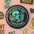 Round enamel pinback button available in 2.5″ or 3″ sizes with a black base and high-gloss finish. The design features neon green and blue gradient skull in the center, pastel pink roses and green leaves below, and mint skeleton hand on the right side. Arched text reads “SILENCE IS NOT SURVIVAL” in teal block letters. Shown styled on a denim jacket, held in hand against bright yellow backdrop, placed in a candy heart flat lay, and propped on pavement in front of graffiti wall.