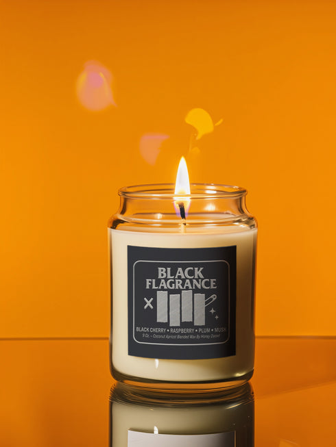 A 9-ounce hand-poured candle in a clear, cylindrical glass jar with a slight taper at the base. The wax is a creamy, solid off-white color, filling the jar to just below the rim where a single white cotton wick is centered. The label features the words "BLACK FLAGRANCE" in a bold, white, sans-serif font. Below this, a stylized graphic mimics a famous punk band logo with four vertical white bars. The candle is topped with a shiny, metallic gold-colored lid.