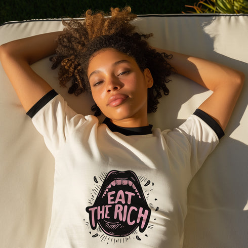 Off-white ringer t-shirt with black contrast trim on the collar and sleeve cuffs. The front features a bold, high-contrast graphic of an open mouth with the phrase "EAT THE RICH" in capital letters, styled in a hand-drawn font. The design is rendered in black and vibrant pink, with expressive line details surrounding the mouth, adding a dynamic, rebellious feel. The t-shirt has a vintage-inspired fit, made from soft cotton fabric with a slightly relaxed silhouette. The black trim enhances the retro aestheti
