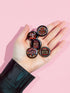 A set of five round, glossy black pinback buttons with embroidered folk art designs and bold activist slogans. Each pin features a different phrase such as "Eat the Rich" and "Resist," surrounded by colorful floral motifs.