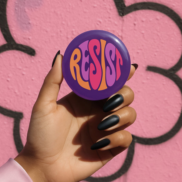 Close-up of a glossy, round purple pinback button. The button features the word "RESIST" in a bold, 70s-style psychedelic font that fills the entire surface. The bubbly, wavy letters alternate in color between a vibrant orange, a light peach-pink, and a deep magenta, creating a powerful and retro statement piece. The design has a distinct groovy and feminist protest aesthetic, making it a perfect accessory for jackets, bags, or activist apparel.