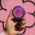 Close-up of a glossy, round purple pinback button. The button features the word "RESIST" in a bold, 70s-style psychedelic font that fills the entire surface. The bubbly, wavy letters alternate in color between a vibrant orange, a light peach-pink, and a deep magenta, creating a powerful and retro statement piece. The design has a distinct groovy and feminist protest aesthetic, making it a perfect accessory for jackets, bags, or activist apparel.