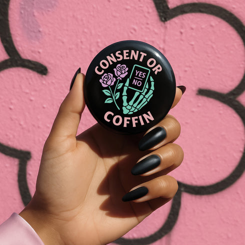 A round black enamel pinback button reading “Consent Or Coffin” in pastel-pink text above and below a mint-green skeleton hand holding a tiny phone interface labeled “YES/NO,” flanked by two stylized pink roses, on a textured purple background.