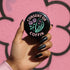 A round black enamel pinback button reading “Consent Or Coffin” in pastel-pink text above and below a mint-green skeleton hand holding a tiny phone interface labeled “YES/NO,” flanked by two stylized pink roses, on a textured purple background.