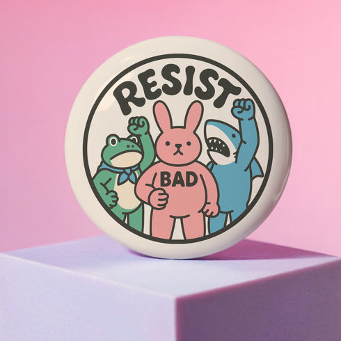 A round white pinback button with the word 'RESIST' in a bold, black, arched font. Below the text stand three cute cartoon characters raising their fists in protest: a green frog with a blue bandana, a pink bunny with '/BAD' on its chest, and a blue shark with sharp teeth. The activist pin has a glossy, protective mylar finish and a durable metal shell, designed as political statement flair for jackets, bags, and backpacks. This punk-inspired, kawaii- badge is a symbol of joyful rebellion.
