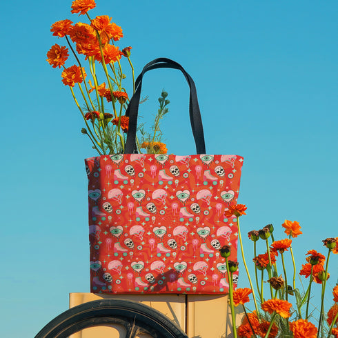 Tote bag with black handles featuring a vibrant red background covered in pastel pink and blue illustrations of skulls, roller skates, eyes, and geometric shapes. Displayed on an orange surface with dramatic shadows.