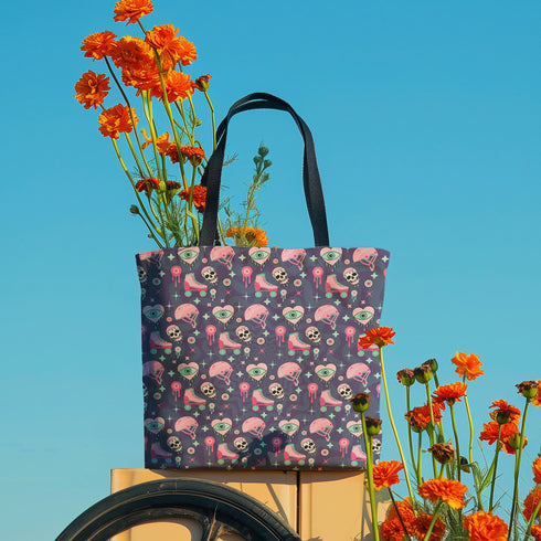 Tote bag with black handles and a purple background featuring a repeating pattern of pink roller skates, white skulls, pink helmets, teal eyeballs, and pink jellyfish-like shapes. The design has a spooky yet playful vibe, combining roller derby elements with supernatural motifs. The overall aesthetic is quirky, retro-inspired, and perfect for skate enthusiasts with a taste for the unconventional.