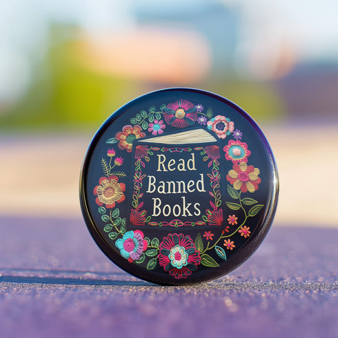 Round black pinback button with "Read Banned Books" in gold lettering on an open book image, surrounded by vibrant embroidery-style flowers in pink, orange, blue and green. Folk art-inspired literary protest pin with glossy finish and safety pin backing, available in 2" and 3" sizes.