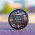 Round black pinback button with "Read Banned Books" in gold lettering on an open book image, surrounded by vibrant embroidery-style flowers in pink, orange, blue and green. Folk art-inspired literary protest pin with glossy finish and safety pin backing, available in 2" and 3" sizes.