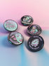 A set of five round 2.25-inch pinback buttons featuring original roller derby designs including a bruised face, saint on skates, flaming roller skate, skull in helmet, and bold text phrases like “Built for Bruises” and “Hell on Wheels.” Each button has a glossy black finish with vibrant pastel pink, purple, mint, and aqua artwork, mounted on metal tinplate with a safety pin back.