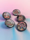 A set of five round, glossy black pinback buttons with embroidered folk art designs and bold activist slogans. Each pin features a different phrase such as "Eat the Rich" and "Resist," surrounded by colorful floral motifs.