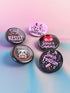 A set of five glossy round pinback buttons with pastel goth and kawaii punk designs, featuring slogans like "Eat The Rich" and "Smash Fascism" in bold pink and black. Each pin has a cute character delivering a powerful message.