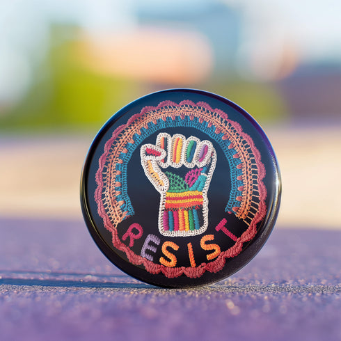 Round pinback metal button with an embroidered-style design on a black background. The center features a raised fist symbol stitched in a multicolored pattern, incorporating rainbow hues and folk-inspired textures. The fist is encircled by a vibrant, lace-like border in shades of blue, orange, and red, resembling intricate embroidery. Below the fist, the word "RESIST" is spelled out in bold, stitched-textured letters in red and orange. The button has a silver metal back with a secure pin closure.