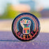 Round pinback metal button with an embroidered-style design on a black background. The center features a raised fist symbol stitched in a multicolored pattern, incorporating rainbow hues and folk-inspired textures. The fist is encircled by a vibrant, lace-like border in shades of blue, orange, and red, resembling intricate embroidery. Below the fist, the word "RESIST" is spelled out in bold, stitched-textured letters in red and orange. The button has a silver metal back with a secure pin closure.