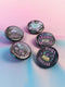 Five glossy 2.25-inch round pinback buttons with pastel punk book-themed designs. Each pin features a different bold phrase like “Read or Die,” “Books Are My Familiar,” and “Give Me Books or Give Me Death,” in pink, purple, and teal colors. Made of tinplate with a safety pin back and glossy scratch-resistant coating.