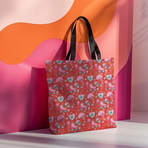 Tote bag with black handles featuring a vibrant red background covered in pastel pink and blue illustrations of skulls, roller skates, eyes, and geometric shapes. Displayed on an orange surface with dramatic shadows.