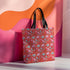 Tote bag with black handles featuring a vibrant red background covered in pastel pink and blue illustrations of skulls, roller skates, eyes, and geometric shapes. Displayed on an orange surface with dramatic shadows.