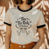 Off-white ringer t-shirt with black contrast trim on the collar and sleeve cuffs. The front features a hand-lettered design with the phrase "Tax the Rich, Feed the Poor, Pet the Cats", arranged in an elegant, whimsical script. Surrounding the text are small decorative elements, including hearts, stars, and delicate floral accents. Below the text, a simple, happy cat illustration with raised paws adds a playful and charming touch. The t-shirt has a vintage-inspired fit, made from soft cotton fabric with a sl