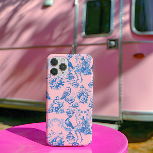 Phone case with pink background featuring blue toile print of skeletons, peacocks, owls, and roses. Victorian-inspired design with gothic elements. Dual-layer protection for various smartphone models.